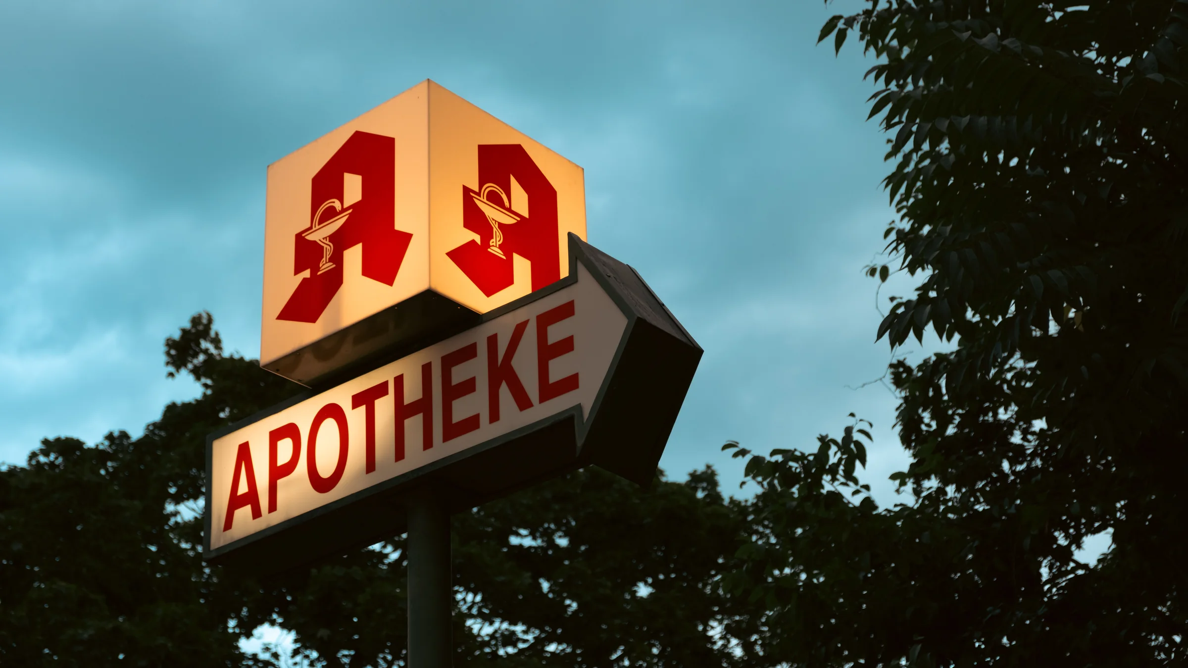 An illuminated German pharmacy sign against a cyan evening sky