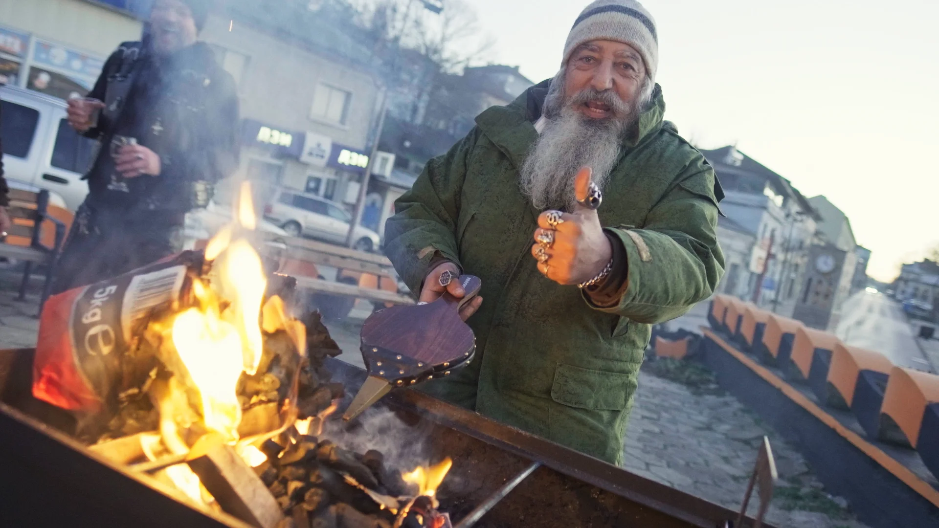 Bearded motorcycle club member giving a thumbs-up beside a fire