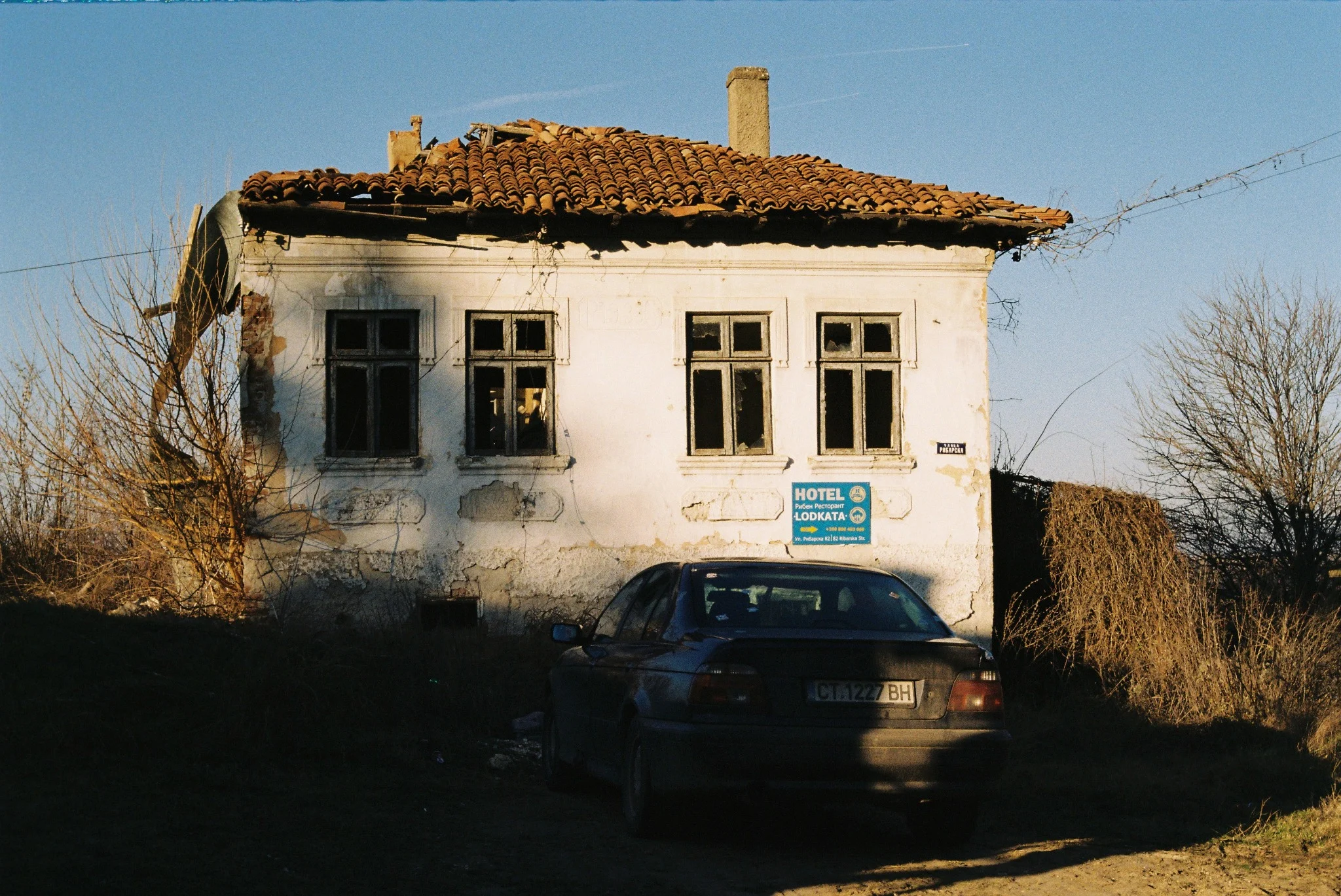 Old roadside hotel with broken windows and a BMW parked in deep winter shadow, photographed on analog film
