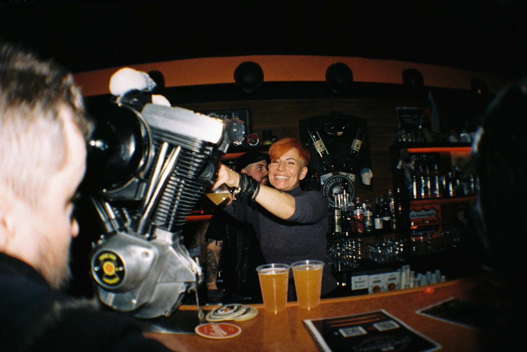 Smiling bartender behind a motorcycle engine and two beers, photographed with direct flash on analog film