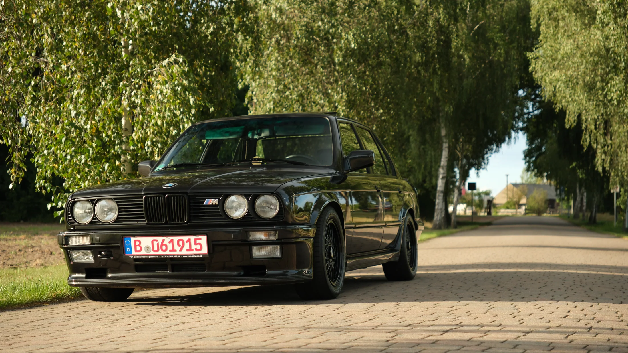 Black classic BMW E30 photographed on a quiet tree-lined road