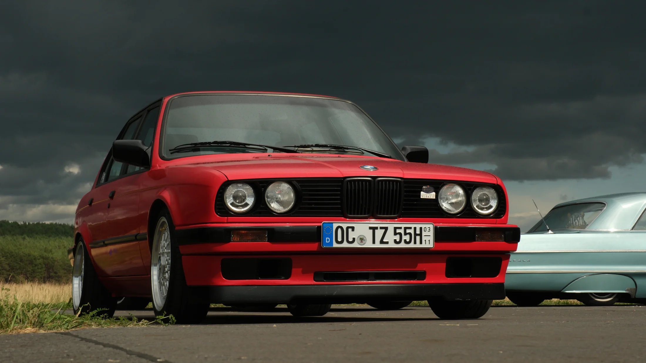 Red classic BMW E30 photographed from a low angle beneath a dramatic storm sky