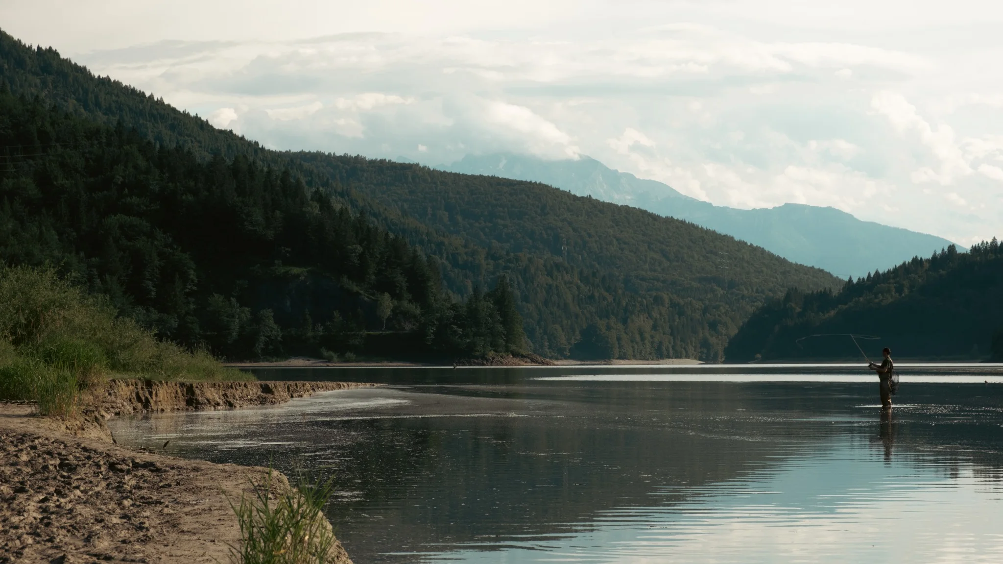 A lone fisherman standing in a mountain lake beneath layered forested ridges