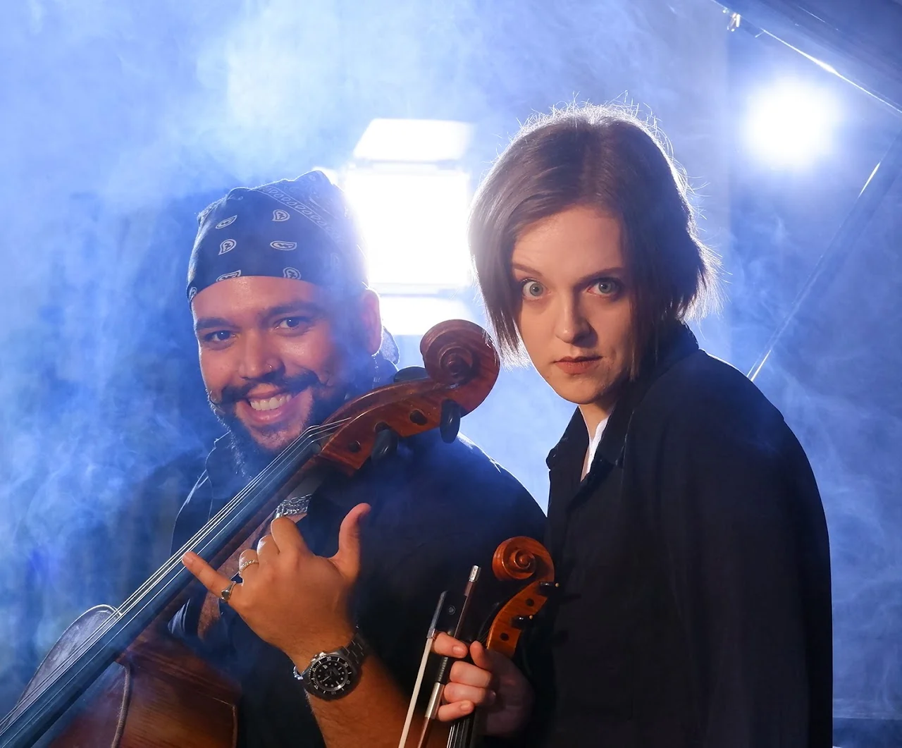 Two string musicians posing together in blue stage smoke